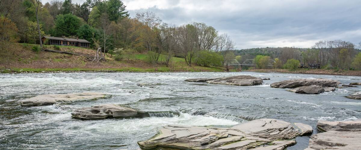 A river with large rocks in the middle, surrounded by trees and a house in the background.