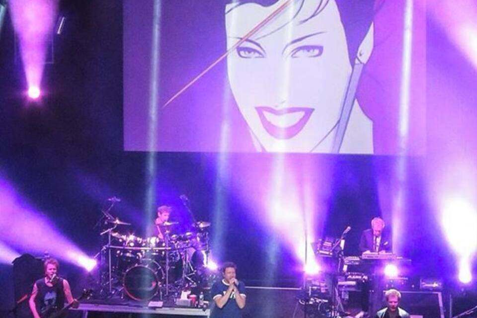 Een band treedt op in de spotlights, met een groot scherm waarop het gezicht van een vrouw wordt getoond.