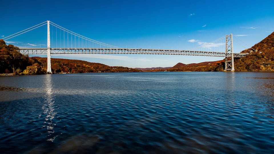 A suspension bridge over a calm river, surrounded by hills and autumn foliage.