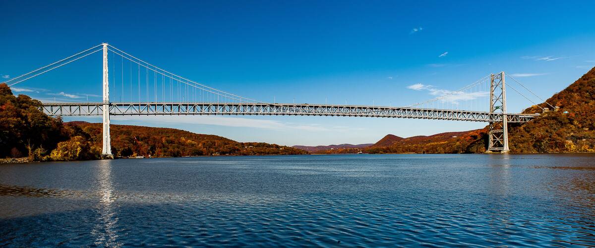 A suspension bridge over a calm river, surrounded by hills and autumn foliage.