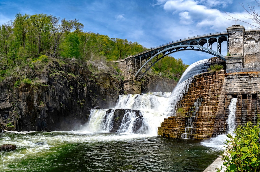 Un barrage en pierre avec une cascade, un pont et une colline boisée.