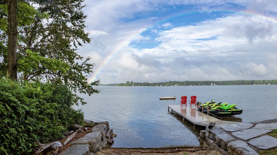A lakeside scene with a rainbow, dock, chairs, and a green jet ski.