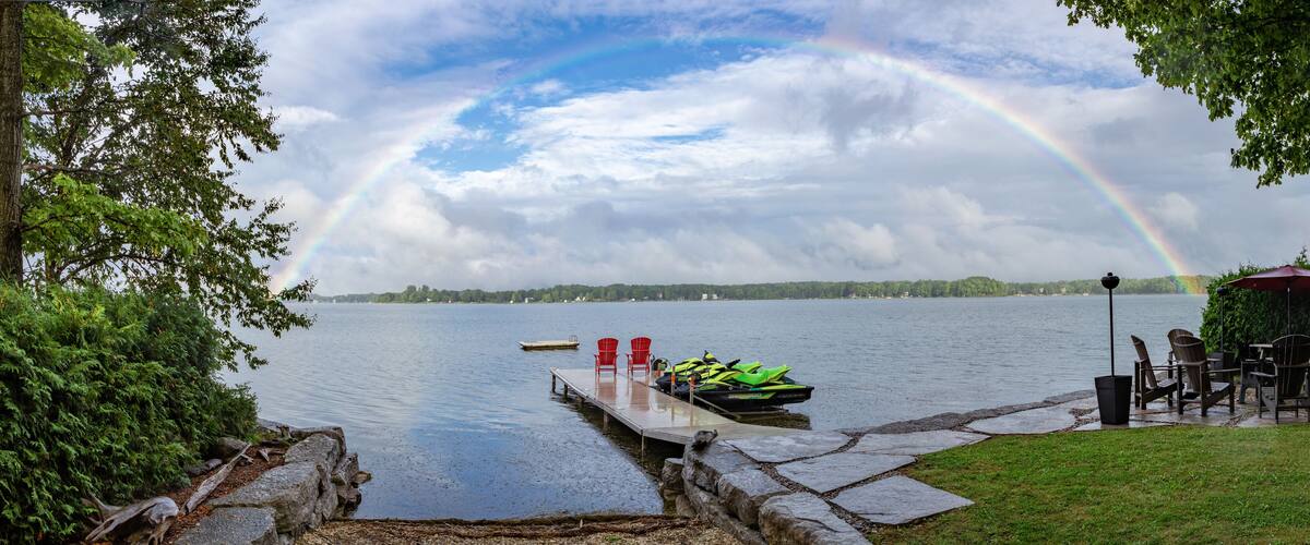 A lakeside scene with a rainbow, dock, chairs, and a green jet ski.