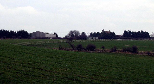 A rural landscape with a large barn, a fenced field, and a small building in the distance.