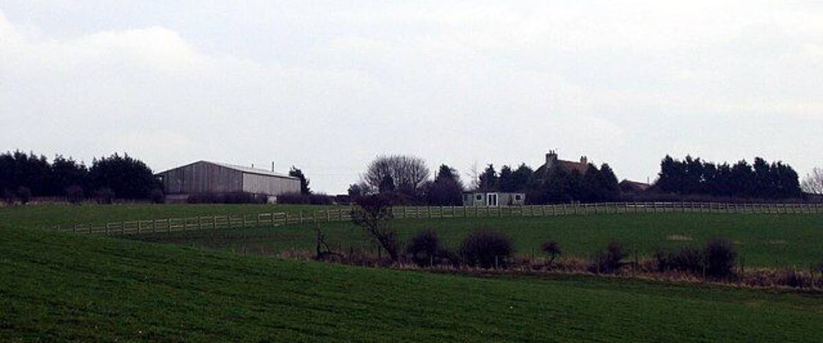 A rural landscape with a large barn, a fenced field, and a small building in the distance.