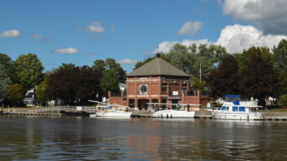 A river with boats docked, a brick building with a dome roof, and trees in the background.
