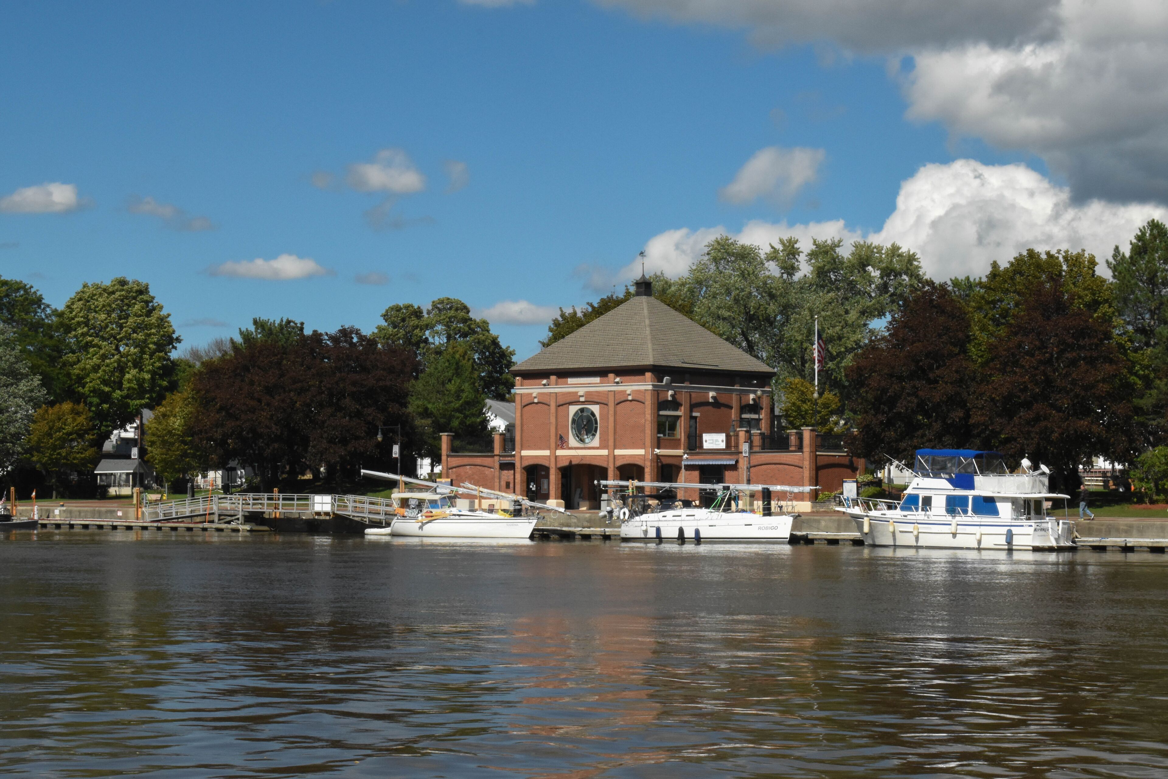 A river with boats docked, a brick building with a dome roof, and trees in the background.