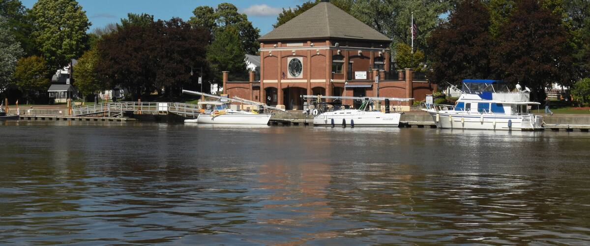 A river with boats docked, a brick building with a dome roof, and trees in the background.