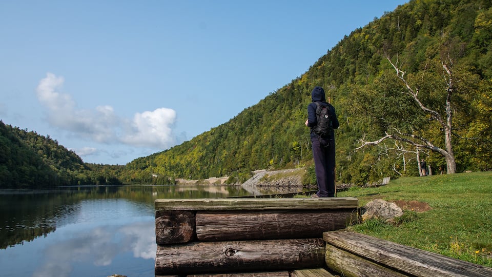 A person standing on a wooden dock by a calm lake, surrounded by lush green hills and trees.