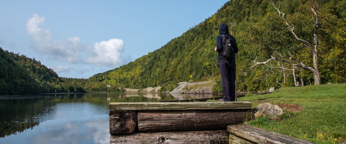 A person standing on a wooden dock by a calm lake, surrounded by lush green hills and trees.