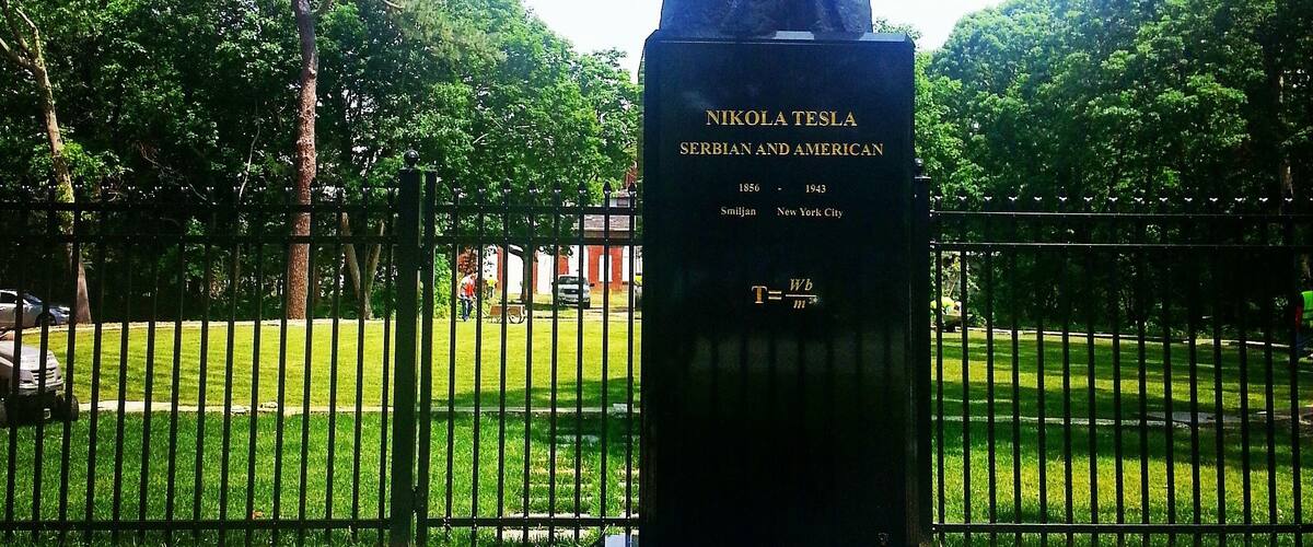 A statue of Nikola Tesla in a park with a plaque, surrounded by trees and a fence.