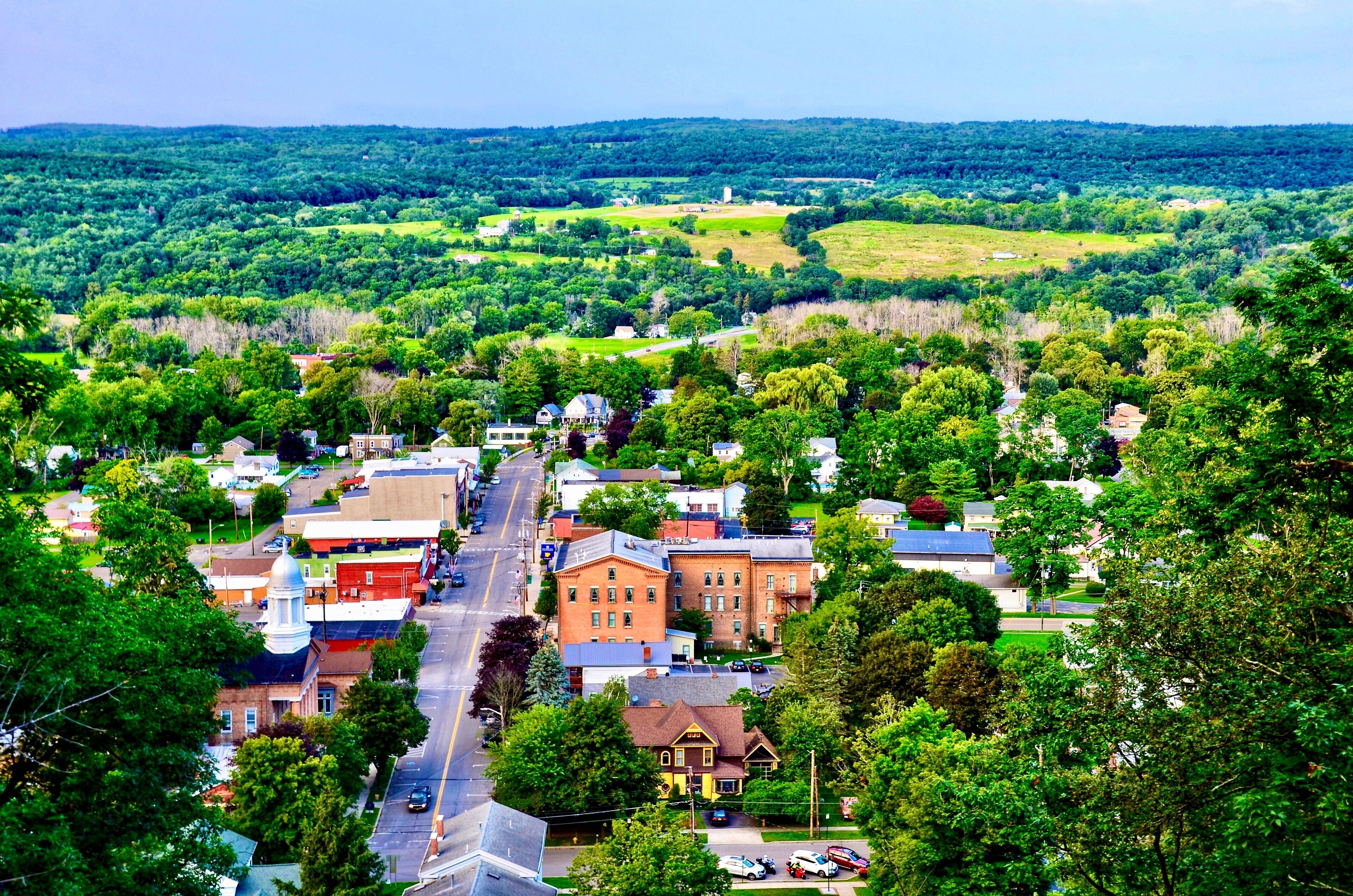 A town with a central street lined with historic buildings, surrounded by lush green hills and a clear sky.