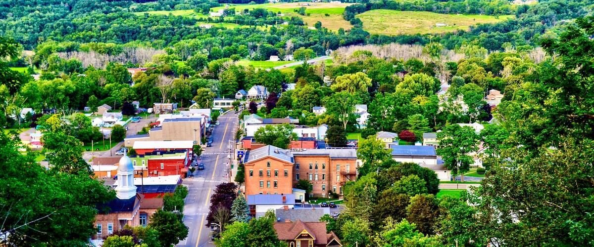 A town with a central street lined with historic buildings, surrounded by lush green hills and a clear sky.
