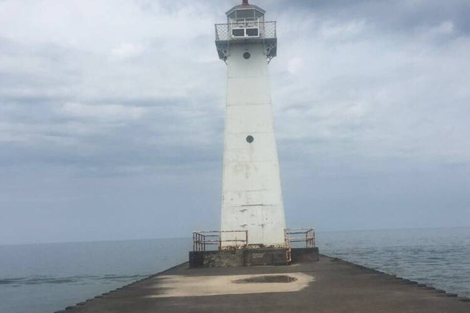 Um farol alto e branco com varanda no topo, localizado em um cais de concreto que se estende para o mar.