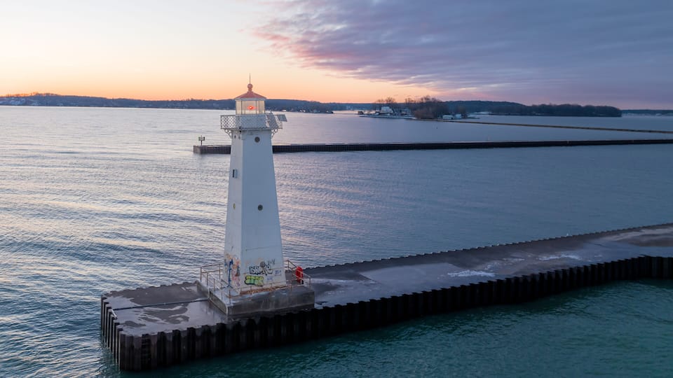 A white lighthouse on a pier extending into a calm body of water, with a sunset sky in the background.