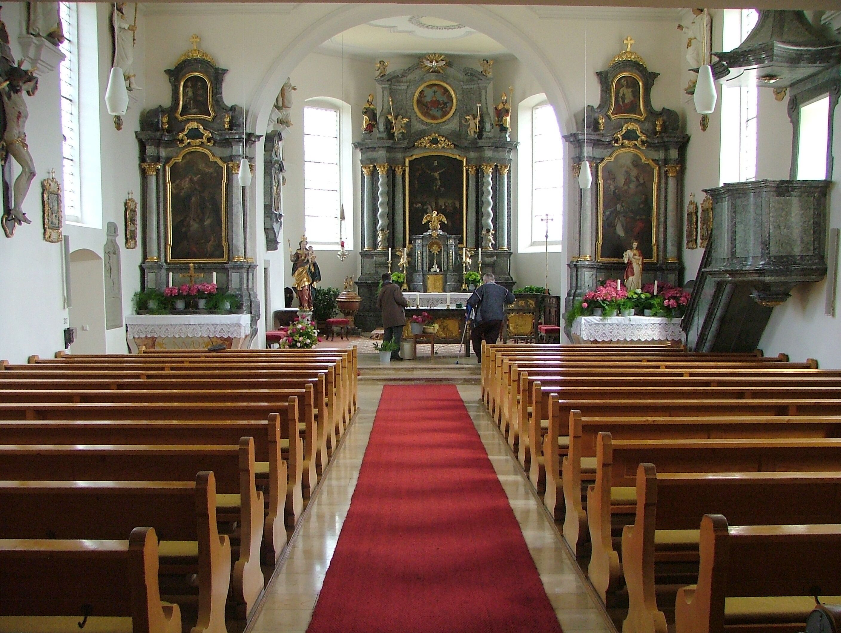 An ornate church interior with wooden pews, a red carpet, and religious statues.