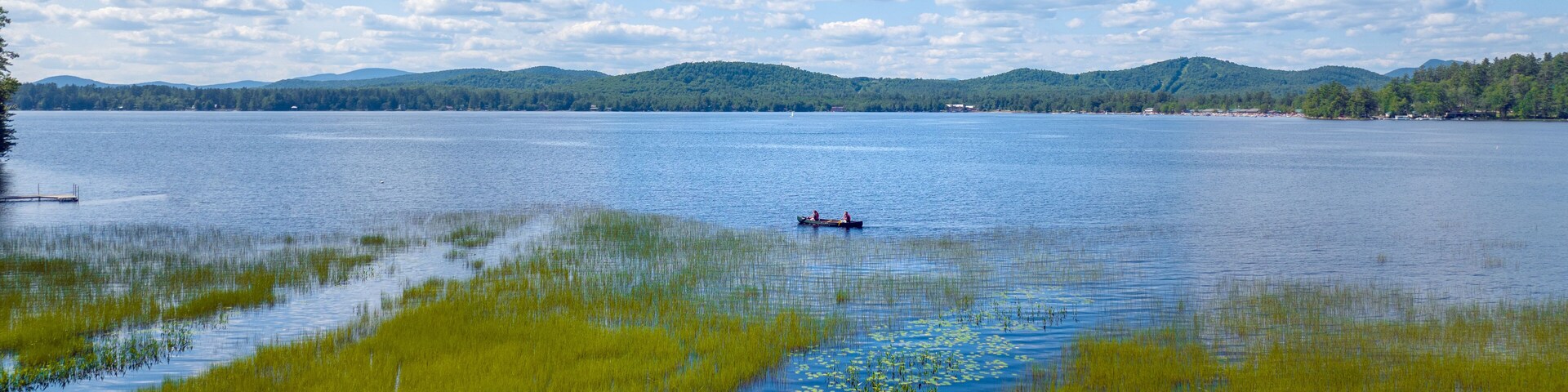 Ein ruhiger See mit einem kleinen Boot in der Mitte, umgeben von üppiger grüner Vegetation und sanften Hügeln im Hintergrund.