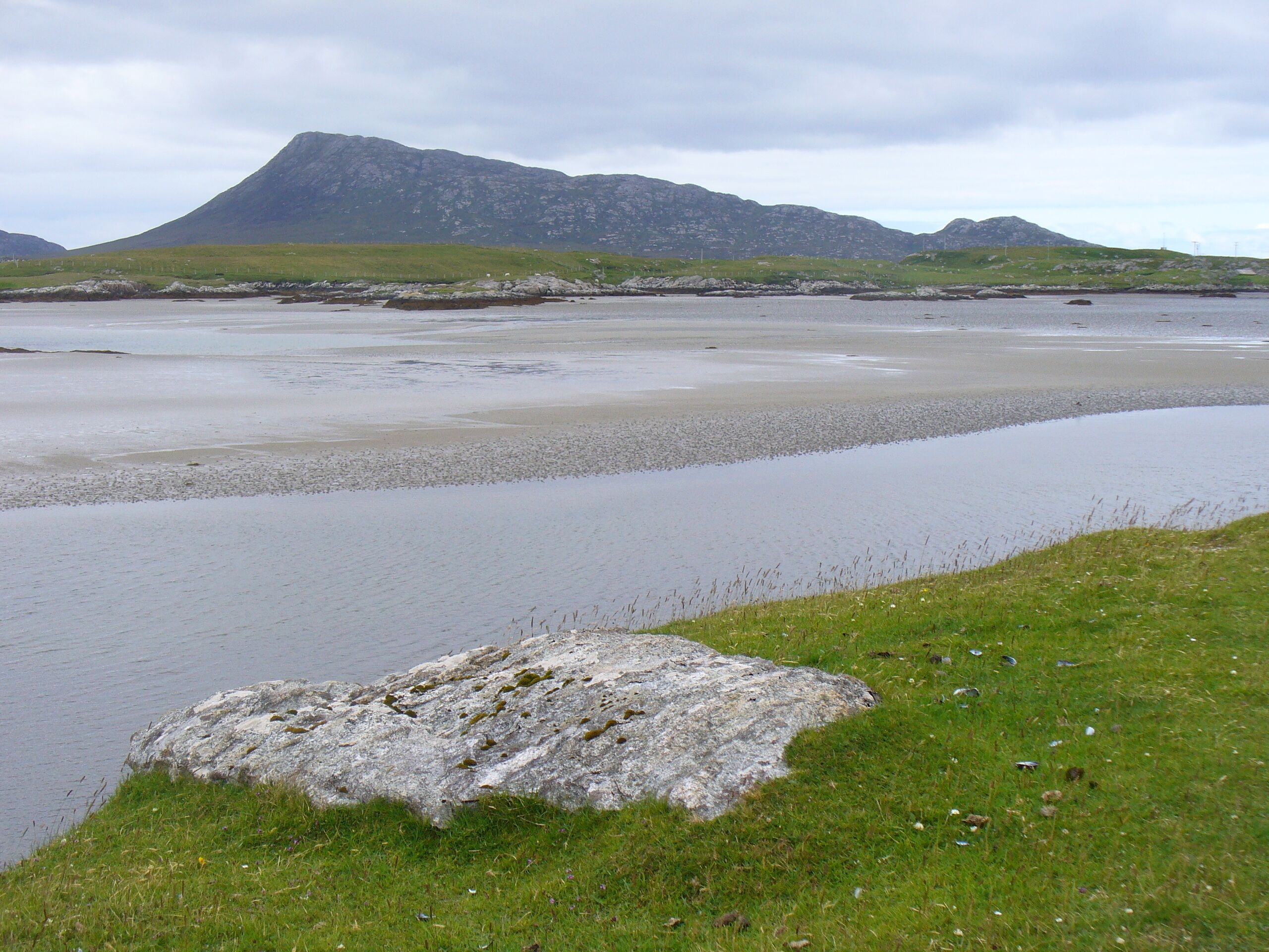A coastal landscape with a rocky outcrop in the foreground, a pebbly beach, and a mountain in the background.