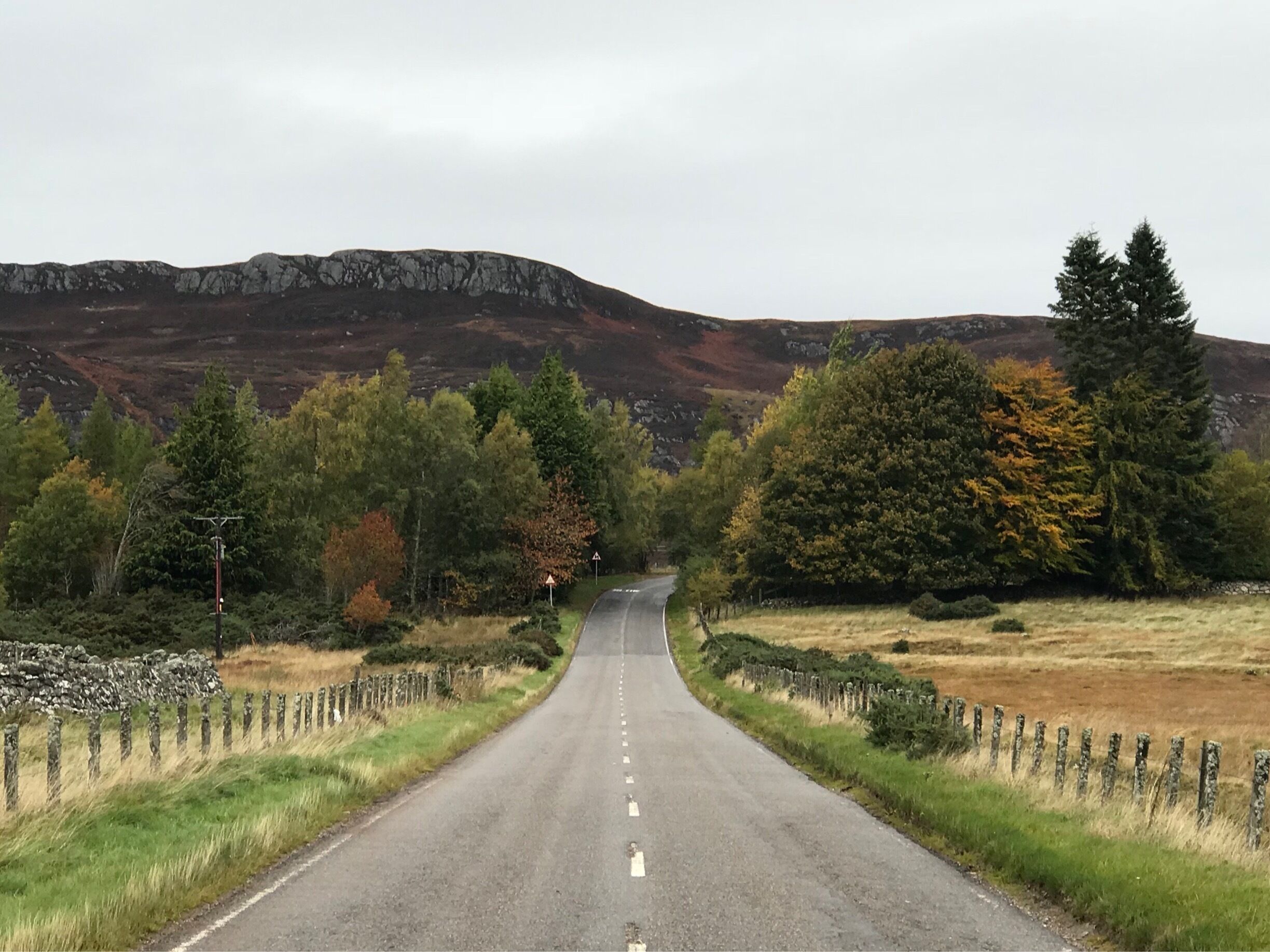 A rural road flanked by stone walls and trees, leading towards a mountainous landscape.