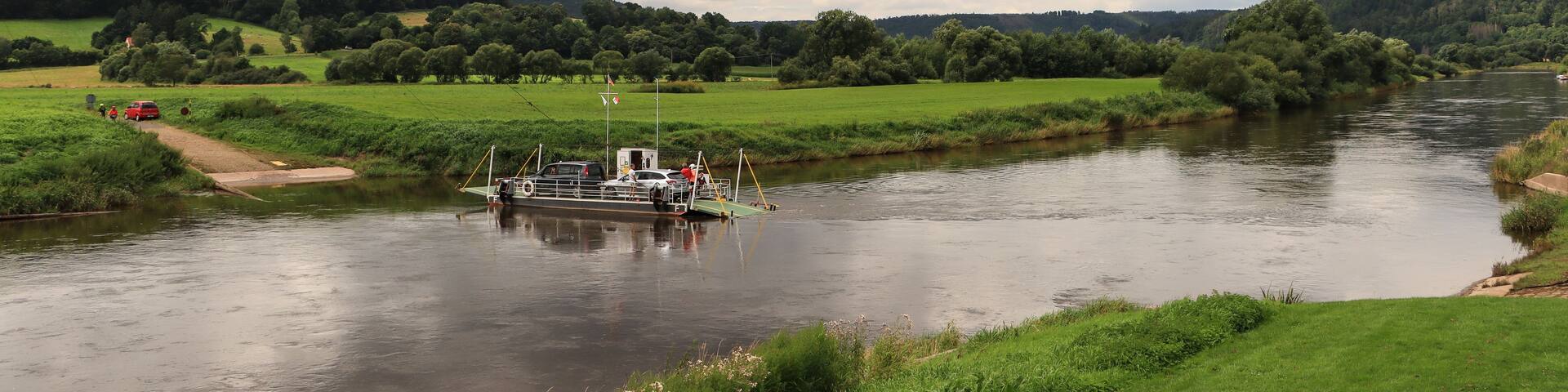 A river with a boat, surrounded by green fields and hills under a cloudy sky.
