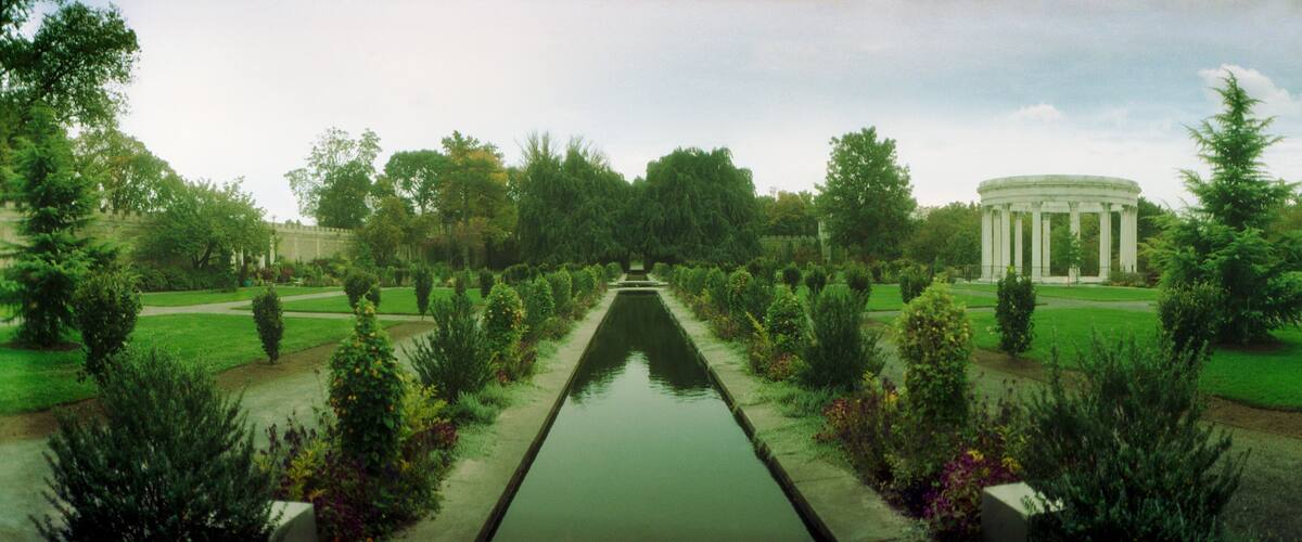 A formal garden with a central water channel, symmetrical landscaping, and a classical building in the background.