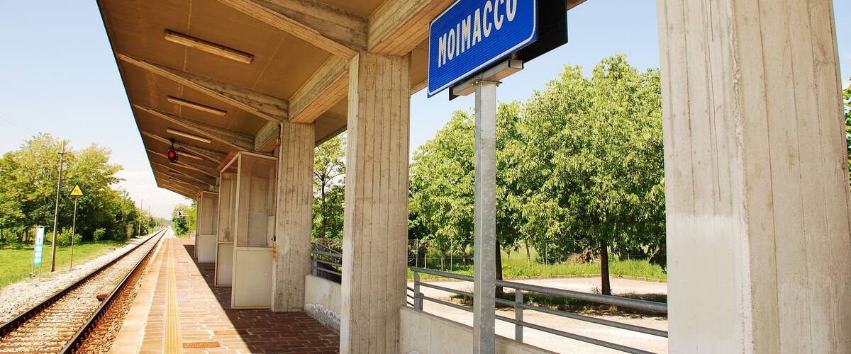 A train station platform with a blue sign reading "MOIMACCO".
