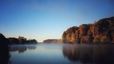 A calm river with mist, surrounded by autumn-colored trees.