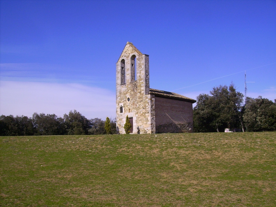 A stone church with a bell tower and a tiled roof, surrounded by a grassy field and trees.