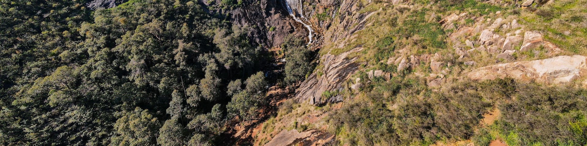 A mountainous landscape with a waterfall, dense forest, and a dirt path.