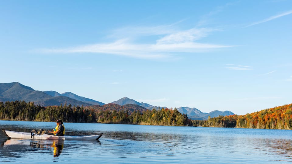 A calm lake with a kayaker, surrounded by autumn-colored trees and mountains in the background.