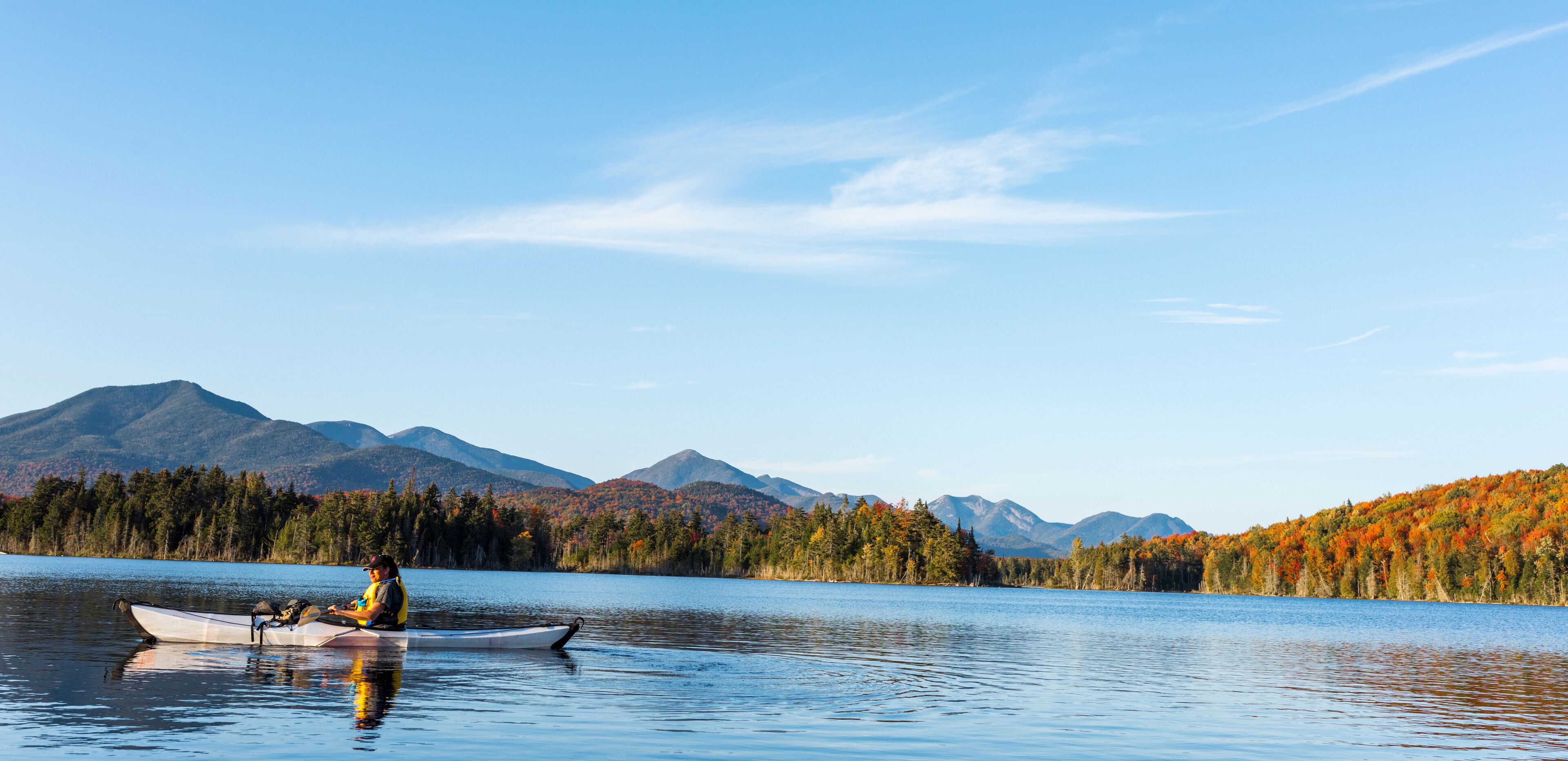 A calm lake with a kayaker, surrounded by autumn-colored trees and mountains in the background.
