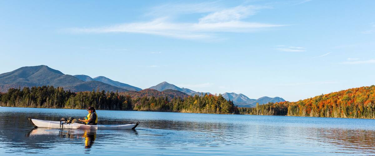 A calm lake with a kayaker, surrounded by autumn-colored trees and mountains in the background.