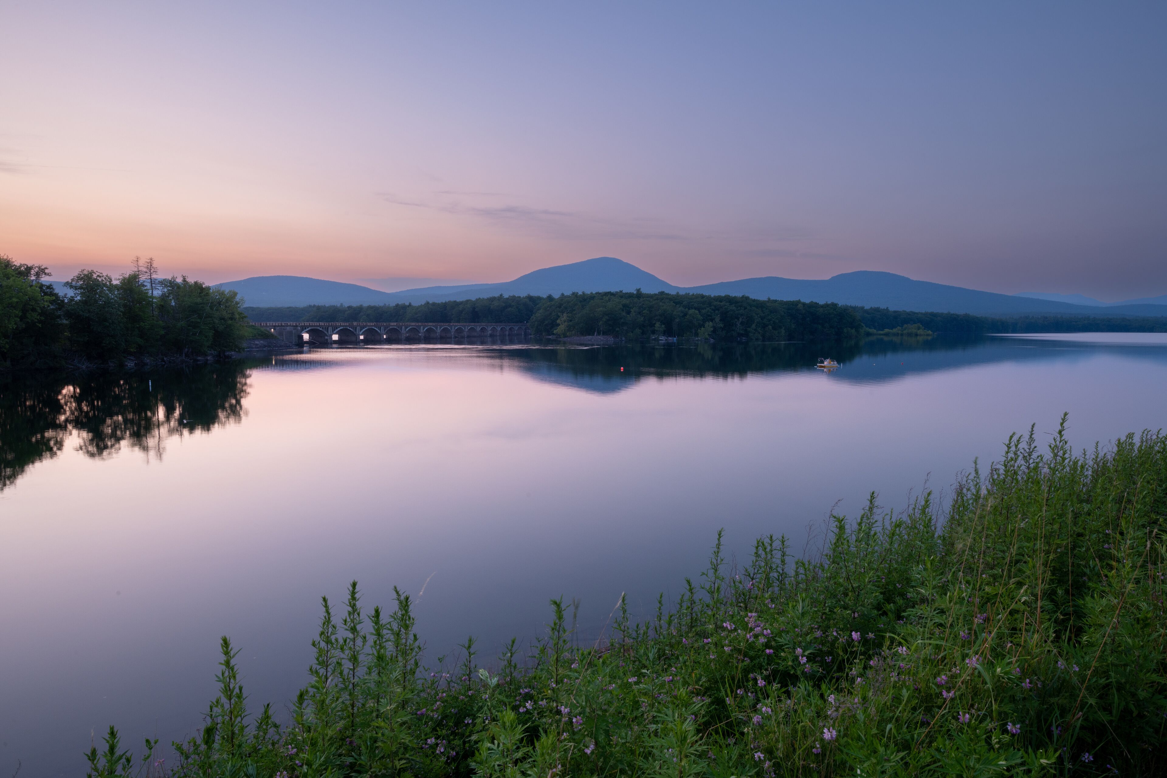A calm lake with a bridge in the distance, surrounded by greenery and mountains.