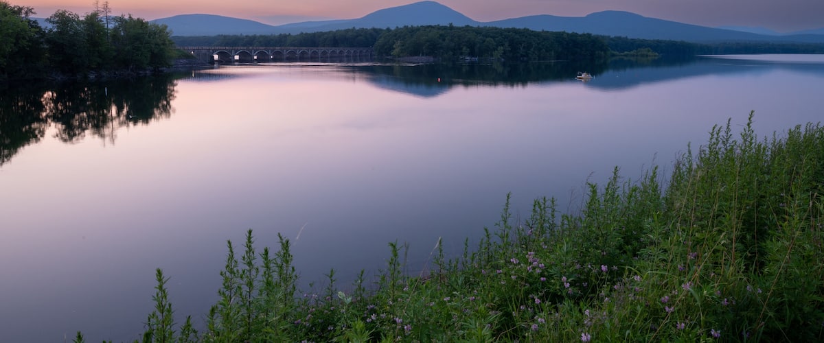 A calm lake with a bridge in the distance, surrounded by greenery and mountains.