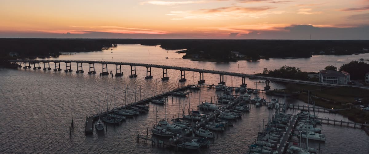 A marina with boats docked, a bridge, and a sunset over the water.