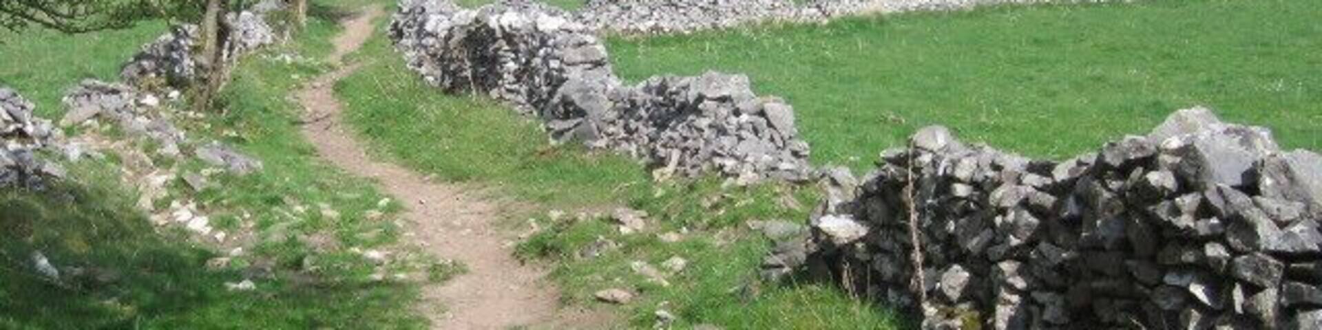 A rural path bordered by stone walls, leading through a green field with distant rolling hills.