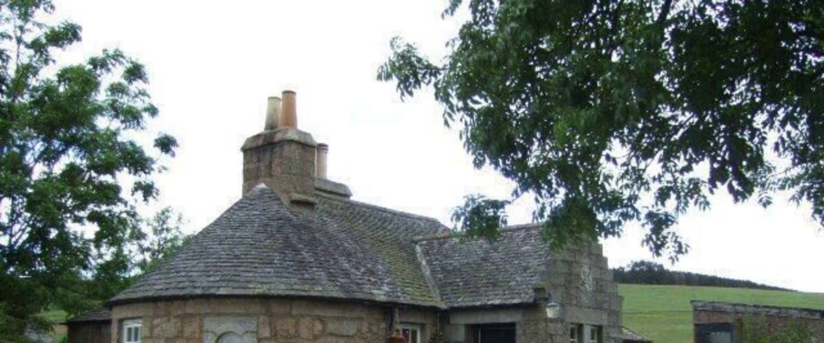 A stone house with a black door and chimney, surrounded by greenery and a gravel driveway.