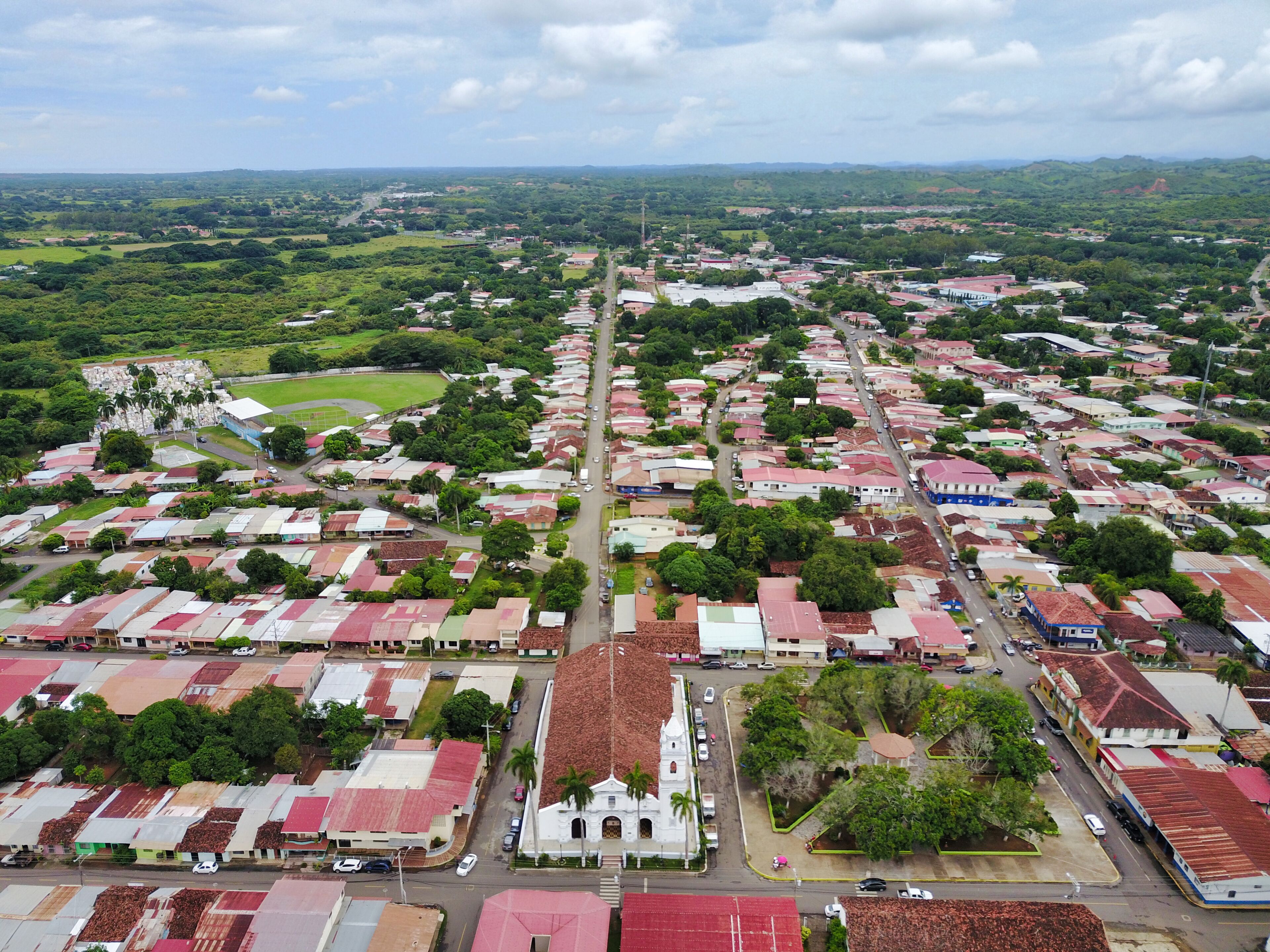 An aerial view of a town with a central road, surrounded by green hills and a prominent white church.