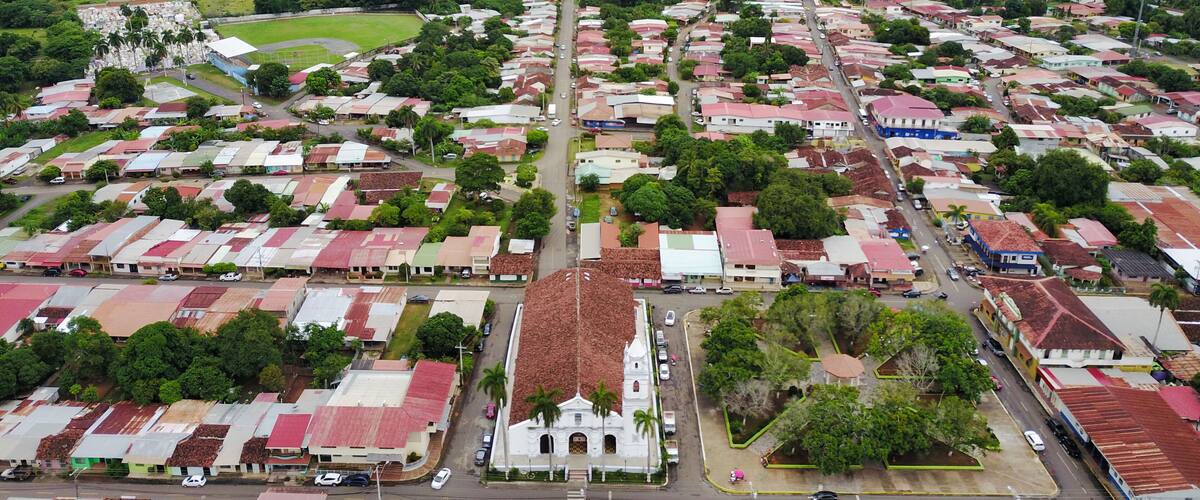 An aerial view of a town with a central road, surrounded by green hills and a prominent white church.