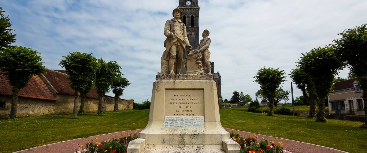 A monument with a clock tower, surrounded by a flower bed and set in a grassy area with trees and buildings in the background.