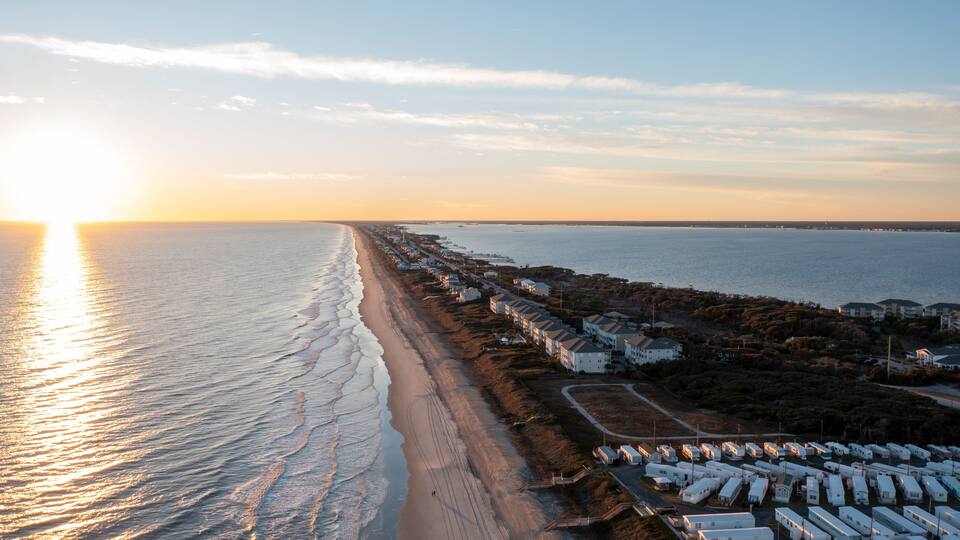 A beach with a row of mobile homes along the shoreline, a sunset in the background, and calm sea waves.