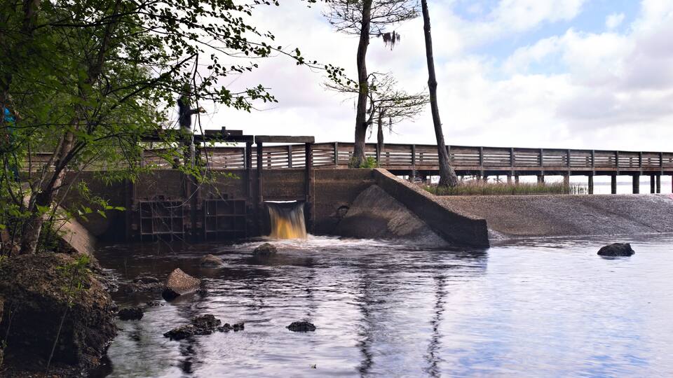 A river flowing under a wooden bridge with a concrete spillway, surrounded by trees and rocks.