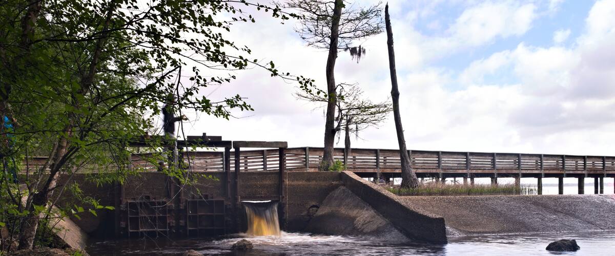 A river flowing under a wooden bridge with a concrete spillway, surrounded by trees and rocks.