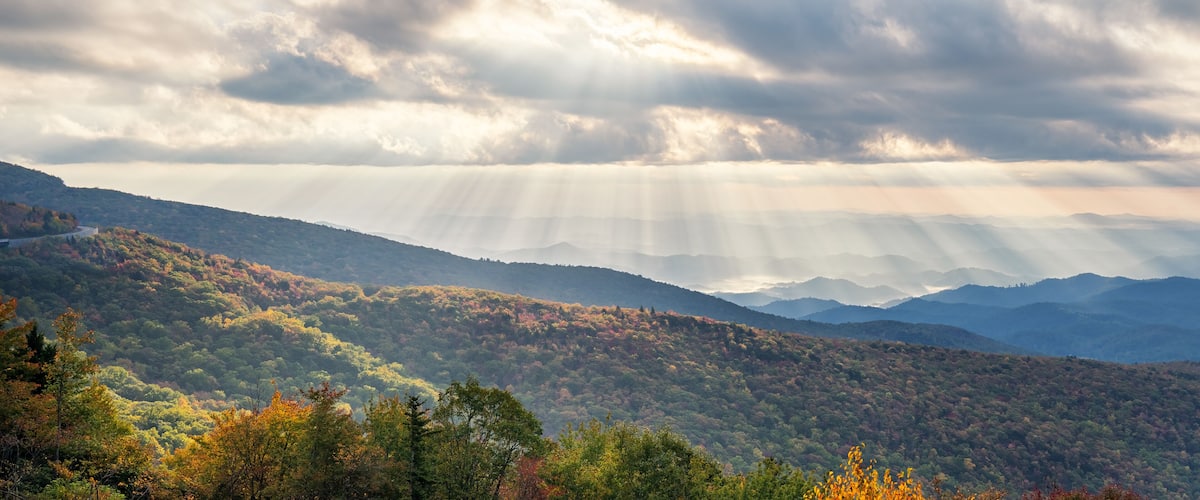 A mountainous landscape with autumn foliage and rays of sunlight piercing through the clouds.