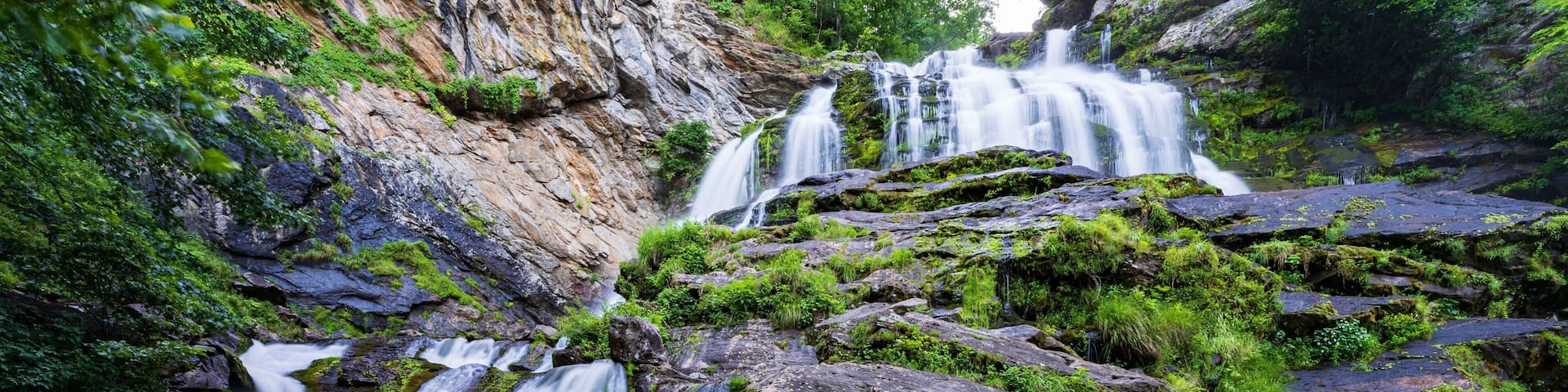 A multi-tiered waterfall cascading over moss-covered rocks in a lush, green forest setting.