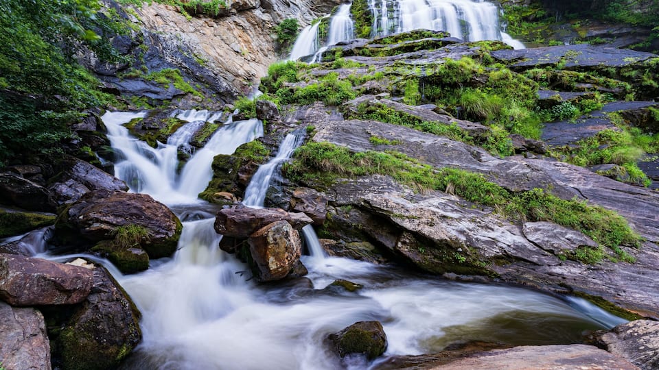 A multi-tiered waterfall cascading over moss-covered rocks in a lush, green forest setting.