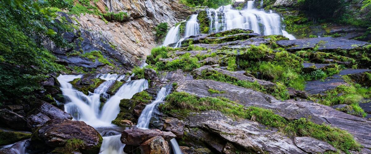 A multi-tiered waterfall cascading over moss-covered rocks in a lush, green forest setting.