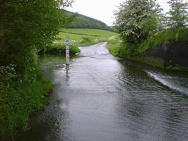 A narrow, wet road with a water crossing and a signpost in a rural, green landscape.