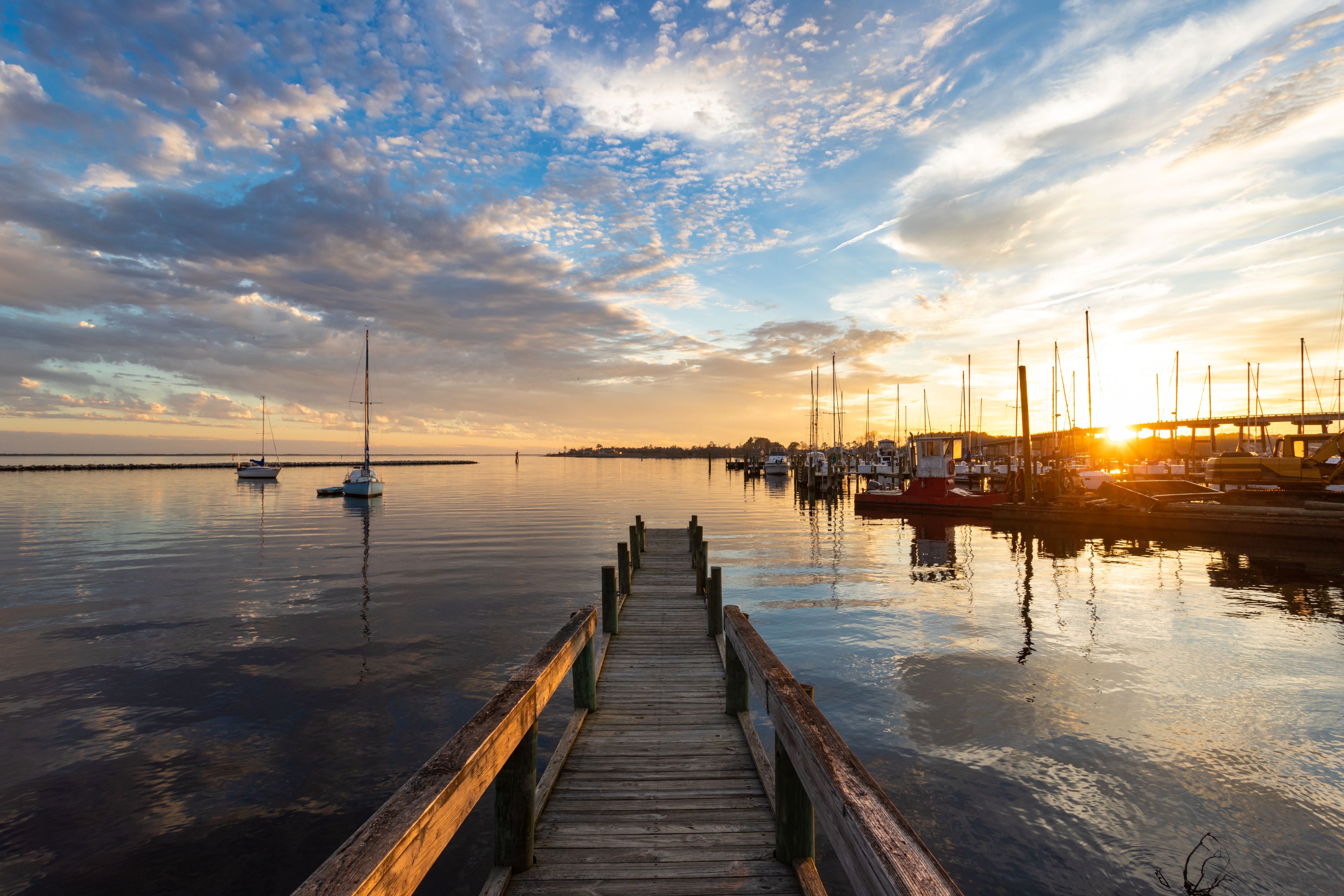 A wooden pier leading to a calm harbor with boats and a bridge in the background.
