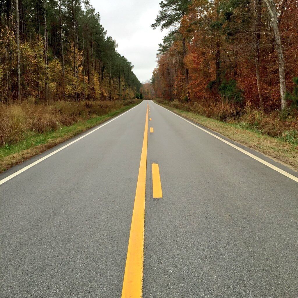 A two-lane road with yellow centerline markings, surrounded by trees with autumn foliage.